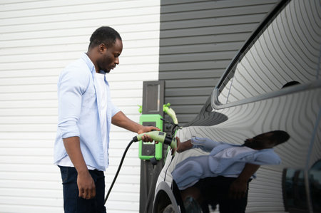 Serious african man holding charge cable in on hand, standing near luxury electric car