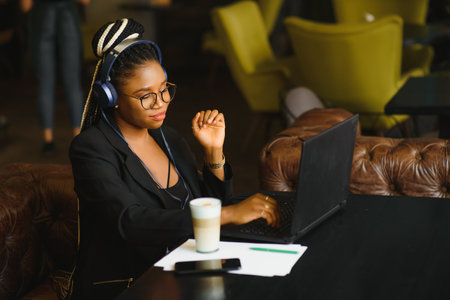 Happy black girl in wireless headphones studying online, using laptop and taking notes, cafe interior, copy spaceの写真素材