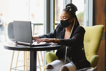 Young african american woman in a protective mask sits in a cafe at a laptop
