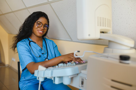 Smiling African woman doctor with ultrasound scanner in hand, working on modern ultrasound scanning machine in light room in clinic. Portrait Of 4D Ultrasound Scanning Machine Operatorの写真素材