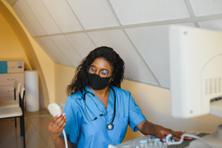 Young cheerful African woman operator in mask of an ultrasound scanning machine analyzing diagnostics results of patient. Young smiling African doctor working on a modern ultrasound equipment.の写真素材
