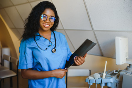 Young afro american female doctor standing in clinic office near modern ultrasound machine.