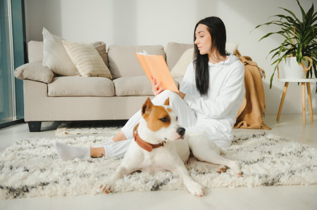 Woman in cozy home clothes relaxing at home with dog reading a book. Comfy lifestyle.