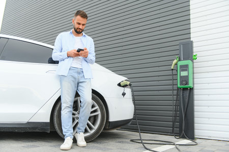 Happy man using smart phone and charging car at vehicle charging station.の写真素材