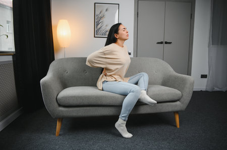 Upset mature woman suffering from backache, unhappy senior blonde sitting on a sofa at living room, feeling because discomfort of pain in back.の写真素材