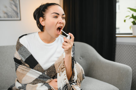 sick young woman in warm clothes spraying throat while sitting on couch at homeの写真素材