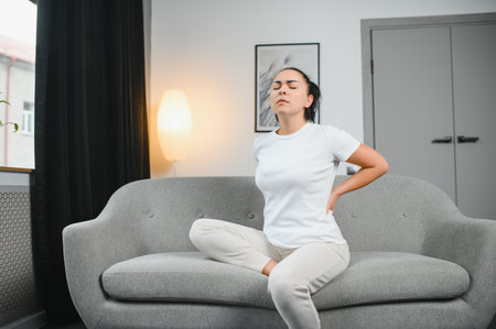 Upset mature woman suffering from backache, unhappy senior blonde sitting on a sofa at living room,...