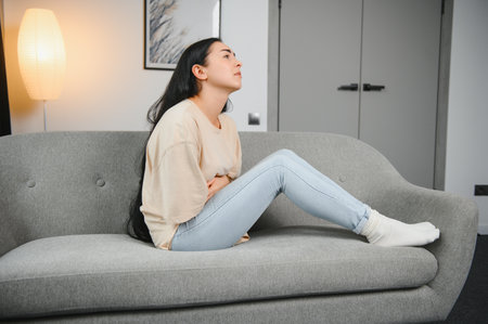 Beautiful hispanic woman in painful expression holding her belly suffering menstrual period pain lying sad on home bed having tummy cramp in female health concept.の写真素材