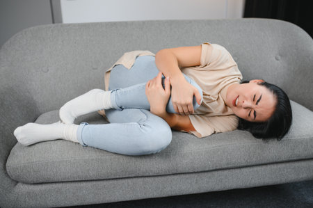 Beautiful hispanic woman in painful expression holding her belly suffering menstrual period pain lying sad on home bed having tummy cramp in female health concept.の写真素材