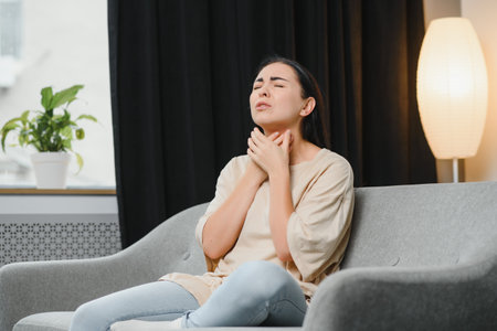 health, cold and people concept - sad sick young Asian woman with sore throat at homeの写真素材