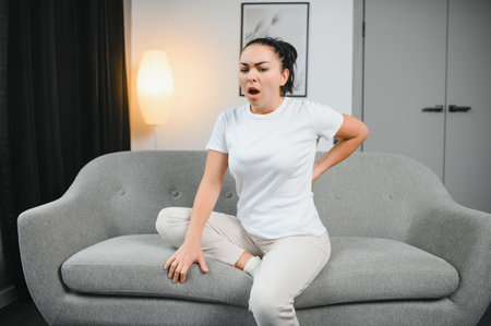 Upset mature woman suffering from backache, unhappy senior blonde sitting on a sofa at living room, feeling because discomfort of pain in back.の写真素材