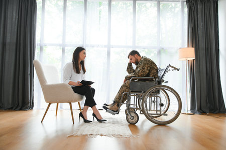 Depressed guy in camouflage uniform paralyzed soldier in wheelchair visiting female psychologistの写真素材