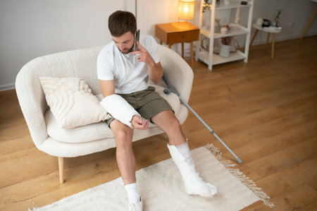 Man with a broken leg and arm using his mobile phone while relaxing on the sofa at home. Accident, injury, treatment, rehabilitation concept.の写真素材