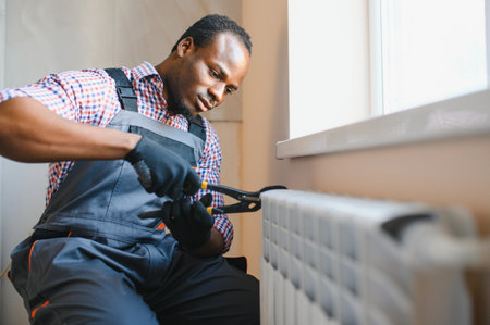 afro repairman in overalls using tools while installing or repairing heating radiator.の写真素材