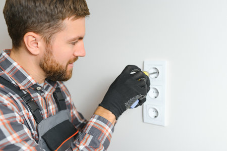 Confident professional electrician in uniform using screwdriver while replacing a socket in apartment after renovation workの写真素材