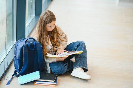 Beautiful schoolgirl smiling while sitting near window and reading book.