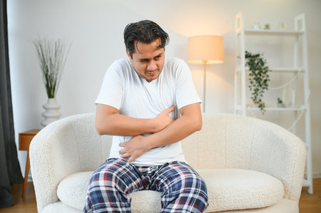 Unhappy Indian or Arabian man, sits on comfortable sofa in cozy living room, holds his hands on his stomach, grimaces from pain in his stomach, suffers from poisoning, spasm, stomach problemsの写真素材