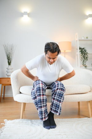 Unhappy Indian or Arabian man, sits on comfortable sofa in cozy living room, holds his hands on his stomach, grimaces from pain in his stomach, suffers from poisoning, spasm, stomach problemsの写真素材