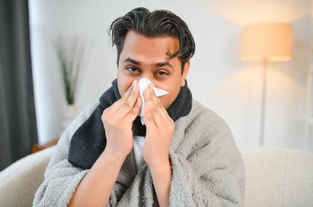 Sick man sitting at home on sofa in living room, sneezing has allergy and runny nose, Indian man...
