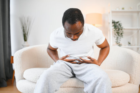 Black Man Having Stomachache Suffering From Painful Abdominal Spasm Standing Touching Aching Abdomen At Home. Abdomen Pain, Stomach Inflammation And Appendicitis Conceptの写真素材