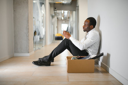 Portrait shot of African American sad businessman in suit and tie sitting with box of stuff. Male office worker lost job. Unemployment rate is growing. Fired upset desperate man.の写真素材