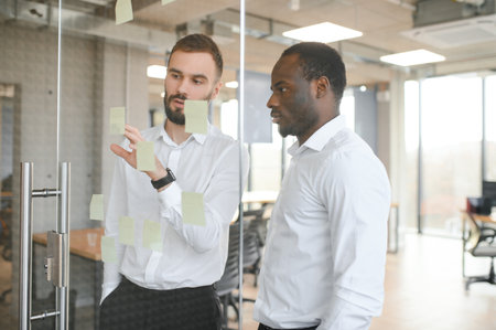 Young Indian businessman manager writing strategy ideas on sticky notes on whiteboard and female African American colleague looking at his strategy scrum presentation. Business project planning.の写真素材