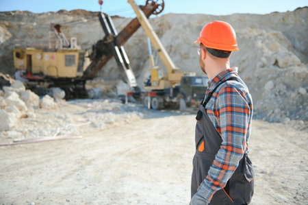 A worker in a helmet stands on the background of an excavator in a quarry.