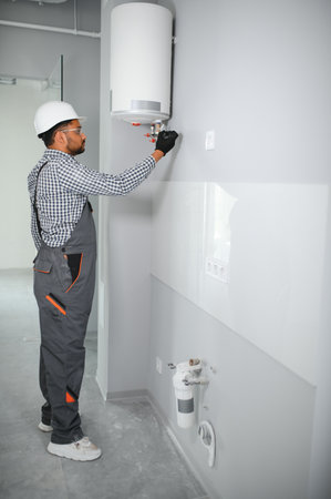 Indian plumber using screwdriver while fixing boiler or water heater, working on heating system in...