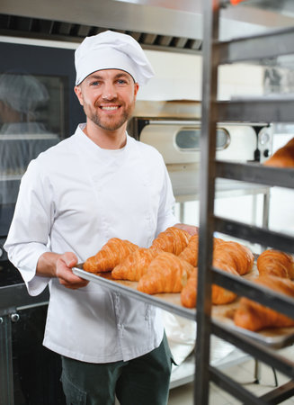 A young handsome male baker holds a tray with French croissants in front of a bakery and smiles.