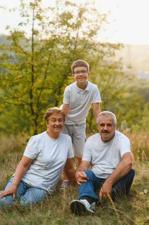 Grandson, grandfather and grandmother sitting on grass having fun outdoors in park at sunset.