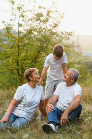 Grandson, grandfather and grandmother sitting on grass having fun outdoors in park at sunset.