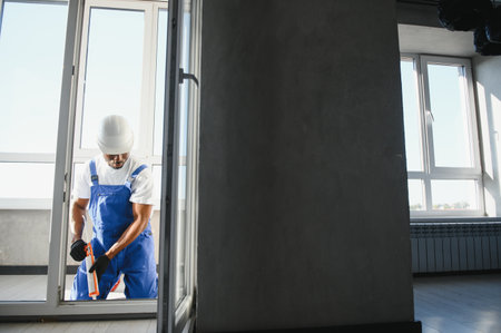 Workman in overalls installing or adjusting plastic windows in the living room at home.