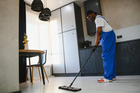 Caution wet floor sign with male young cleaner mopping empty room or office in background. Man janitor with mop in hands and bucket from cleaning service washing gray floor in uniform.の写真素材