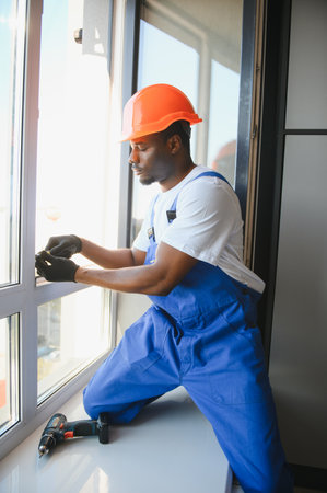 Young African Repairman In Overalls Installing Window.
