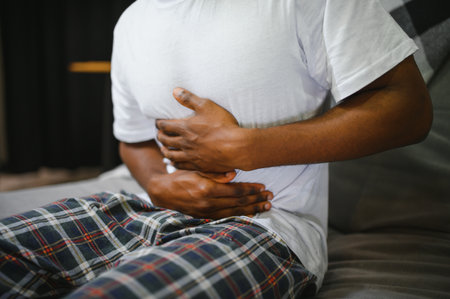 Man clutching his stomach and expressing discomfort while sitting on a sofa in a modern living room. Potential scene related to health issues or digestive problems.の写真素材