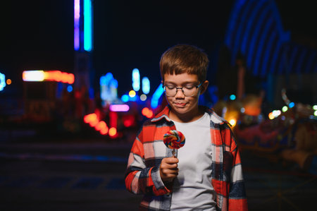 A child enjoying an evening at an amusement park, holding a lollipop and wearing glasses with a plaid shirtの写真素材