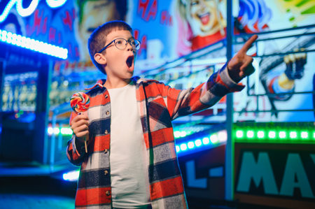 A child at an amusement park showing excitement and joy, holding a lollipop while pointing at something. The background features colorful neon lights and carnival decorations.の写真素材