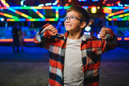 A child enjoying time at an amusement park in the evening, wearing glasses and a plaid shirt, with colorful lights in the backgroundの写真素材