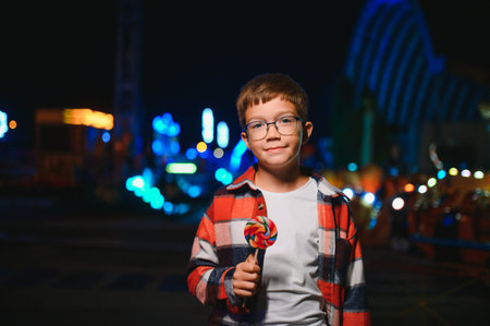 A child eating a colorful lollipop standing against the background of an amusement park with blue lights in the evening.の写真素材