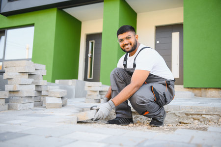 Indian master in gloves lays paving stones in layers. Garden brick pathway paving by professional paver worker. Laying gray concrete paving slabs in house courtyard on sand foundation base.の写真素材