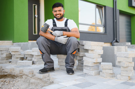 Indian master in gloves lays paving stones in layers. Garden brick pathway paving by professional paver worker. Laying gray concrete paving slabs in house courtyard on sand foundation base.の写真素材