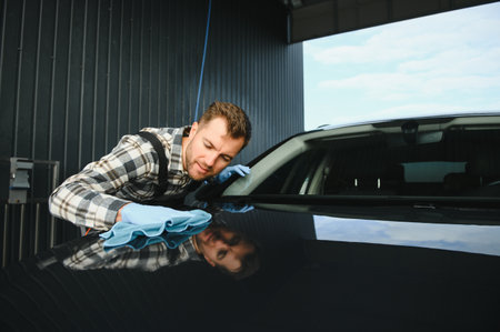 Car detailing, washing and cleaning concept. Young bearded male professional car wash worker in black rubber gloves, holding microfiber and wiping body of luxury car.の写真素材