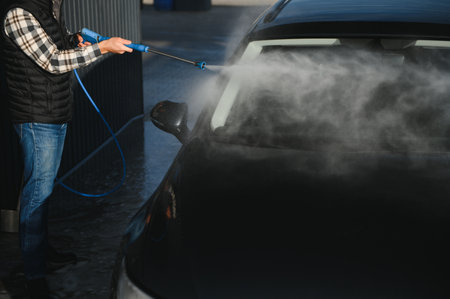 A man washes his car at a self-service car wash using a hose with pressurized water.の写真素材
