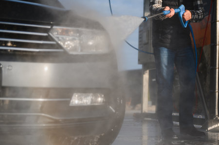 A man washes his car at a self-service car wash using a hose with pressurized water.の写真素材