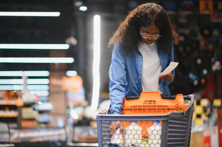 African American Female Customer Posing With Shop Cart Buying Food Products In Supermarket, Smiling Looking At Camera. Black Woman Choosing Groceries In Store Conceptの写真素材