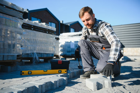 The master in gloves lays paving stones in layers. Garden brick pathway paving by professional paver worker. Laying gray concrete paving slabs in house courtyard on sand foundation base.の写真素材