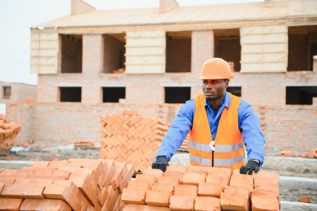 An African American construction worker, in work clothes and a hard hat, is building a brick house.