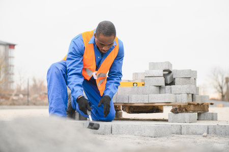 African American master in gloves lays paving stones in layers. Laying gray concrete paving slabs in...