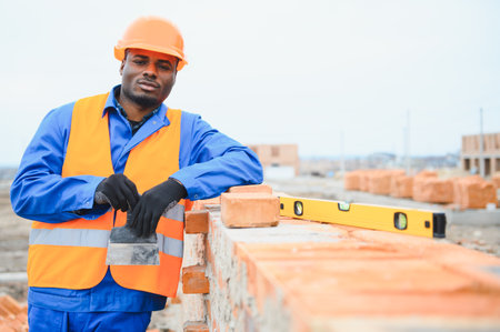An African American construction worker, in work clothes and a hard hat, is building a brick house.の写真素材