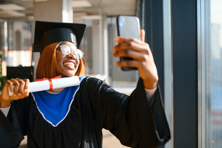 Happy african american female student holding university diploma while taking selfie after graduation ceremony.の写真素材